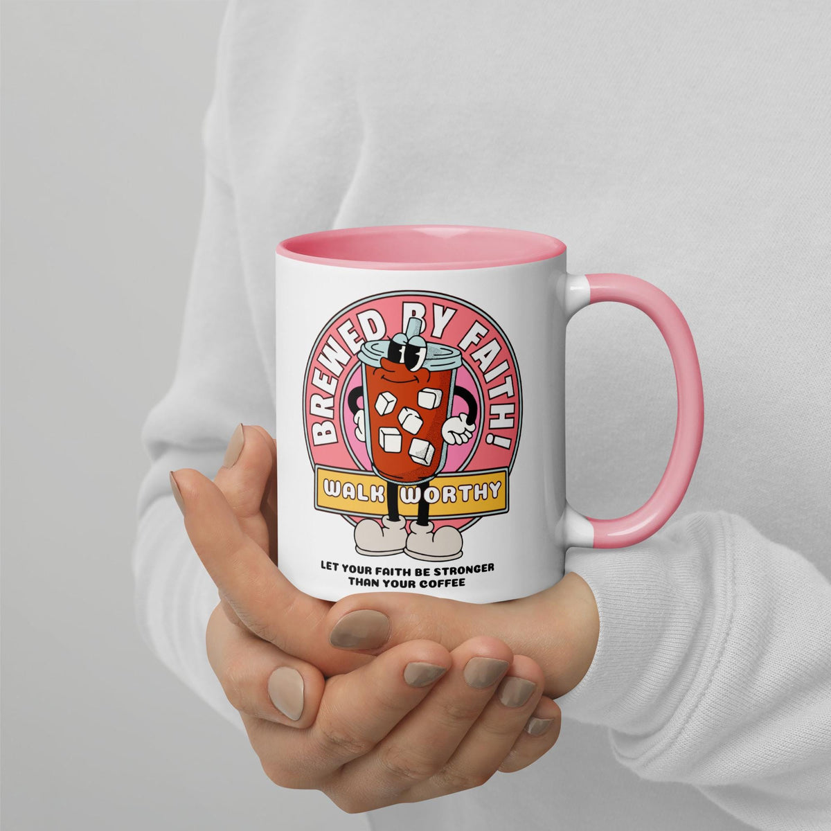 white coffee mug with a pink handle and interior. The mug features a cartoon graphic of an iced coffee drink with sunglasses and a smiling face. The graphic is surrounded by a pink circle with the words 'Brewed by Faith!' and 'Walk Worthy'. Below the graphic is the text 'Let your faith be stronger than your coffee'.