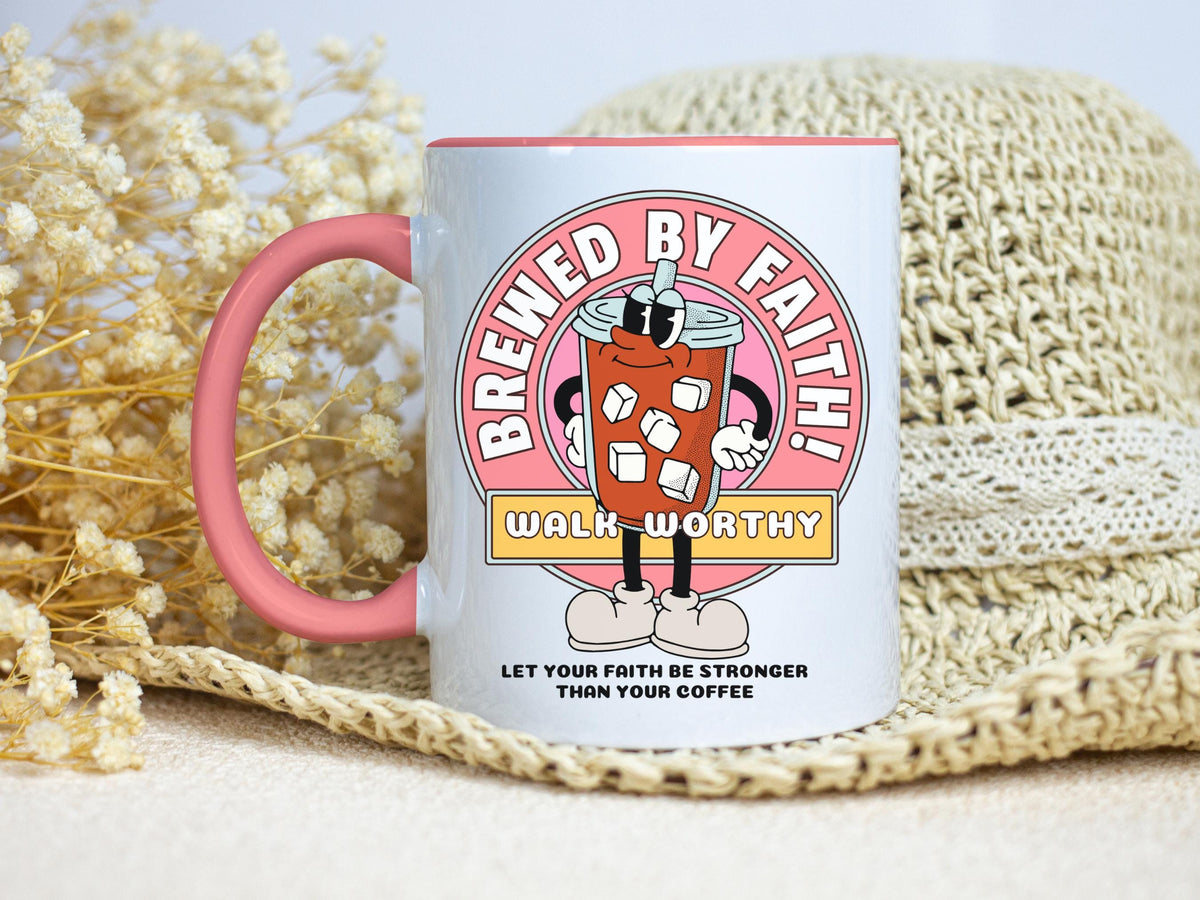 white coffee mug with a pink handle and rim. The mug features a cartoon graphic of an iced coffee drink with the words 'Brewed by Faith!' and 'Walk Worthy' in a circular design. Below the graphic is the text 'Let your faith be stronger than your coffee.'