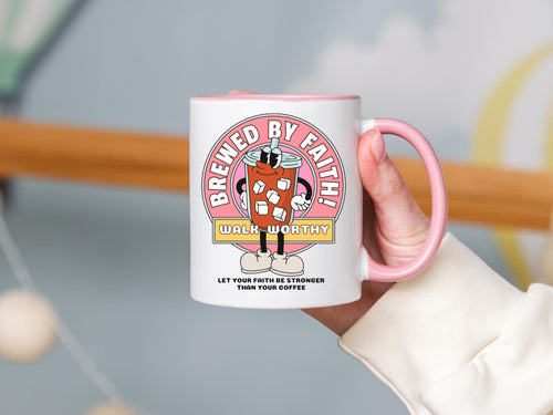 A white coffee mug with a pink handle and rim. The mug features a cartoon graphic of an iced coffee drink with a straw and ice cubes. The graphic is inside a pink circle with the words 'BREWED BY FAITH!' The words 'WALK WORTHY' and 'LET YOUR FAITH BE STRONGER THAN YOUR COFFEE' are also on the mug.